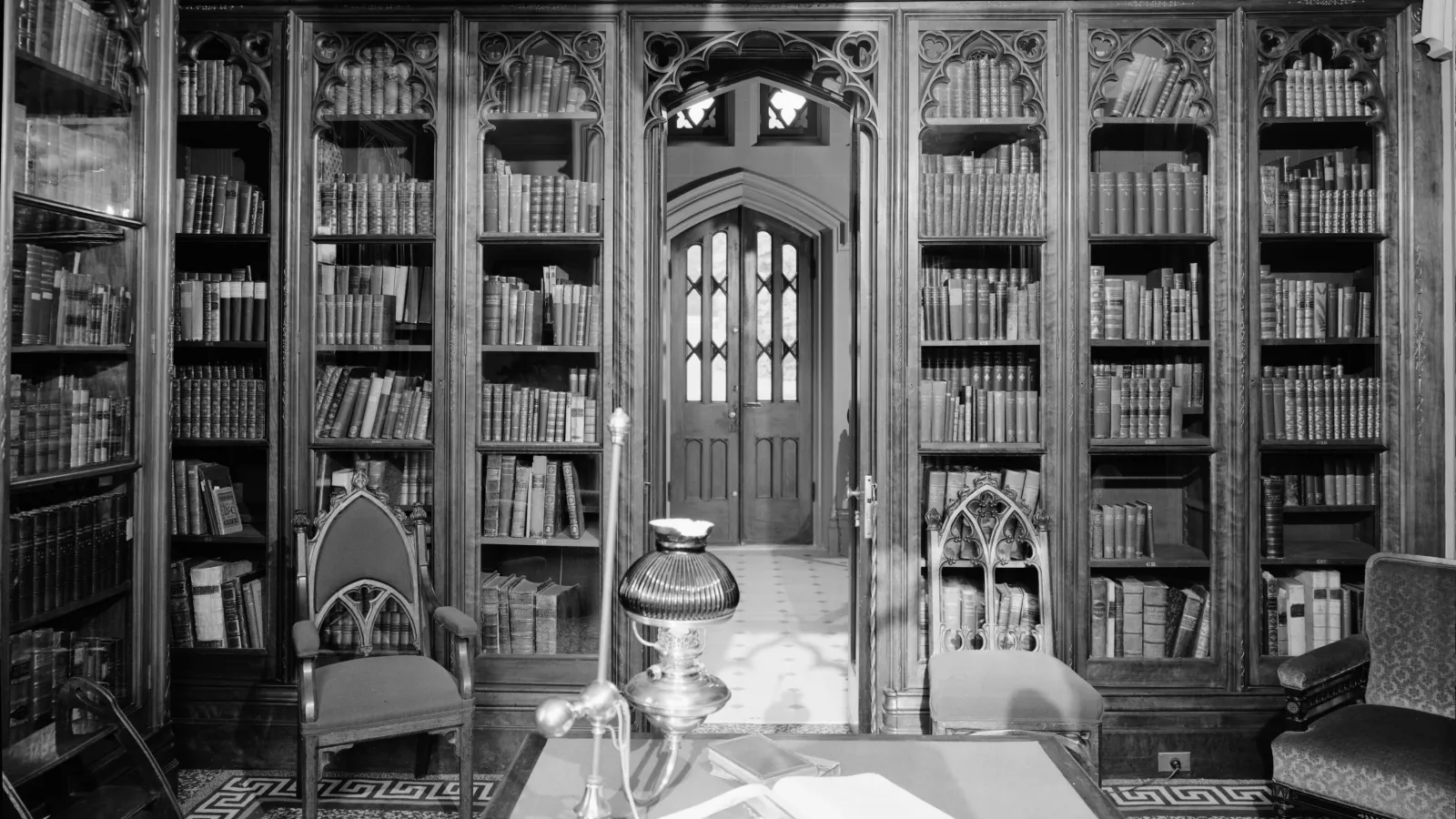 A gothic-revival library wall: glass-fronted bookcases filled with bound volumes, with an open book on a table in front of them.