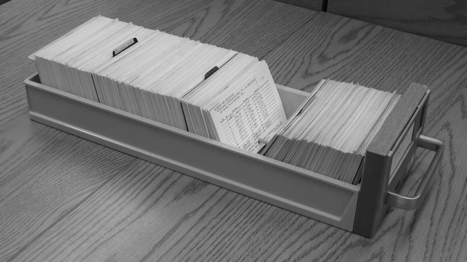 A long library card catalog drawer open on an oak table, packed with index cards, one card pulled up showing handwritten entries.